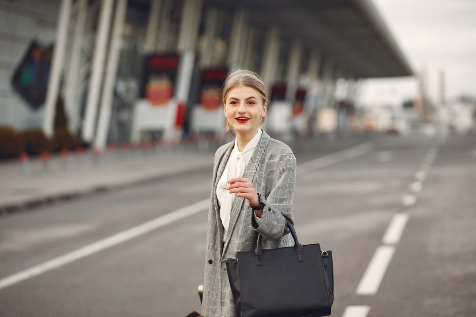 Positive female passenger wearing formal plaid suit standing on asphalt road to airport with handbag and suitcase while smiling looking at camera during business trip