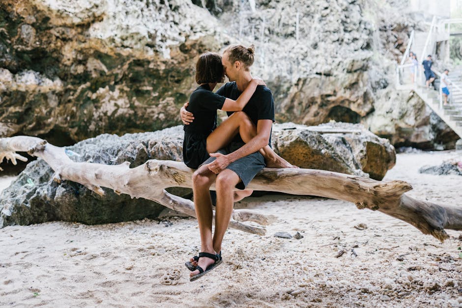 A couple shares an intimate moment on driftwood at a scenic beach location.