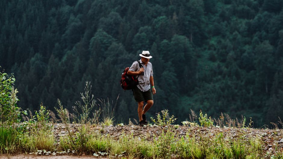 A man in a hat and backpack hiking along a lush green mountain path.