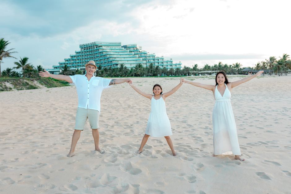 A joyful family holding hands on a sandy beach in Hội An, with a resort and palm trees in the background.