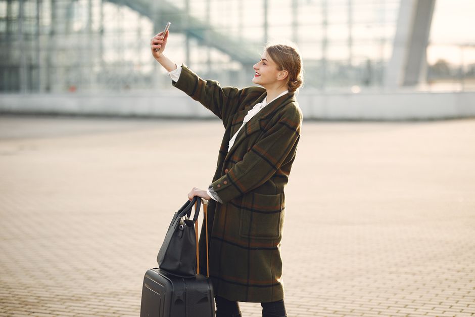 Side view of smiling cheerful female traveler with suitcase and bag wearing trendy plaid coat using smartphone for selfie while standing against contemporary building on city street