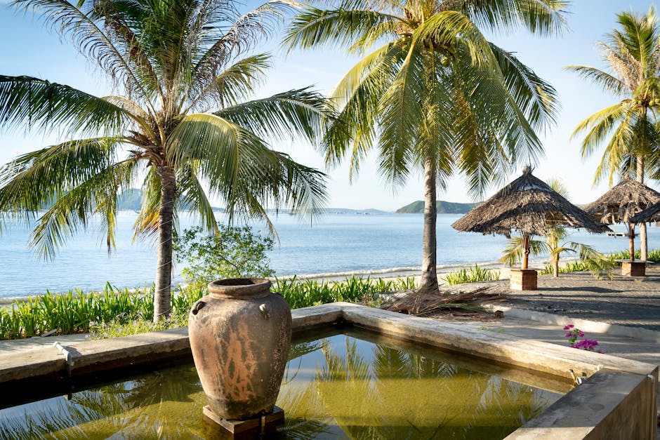 Small decorative fountain basin near exotic lush palms and umbrellas on sandy seacoast in tropical resort on clear sunny day