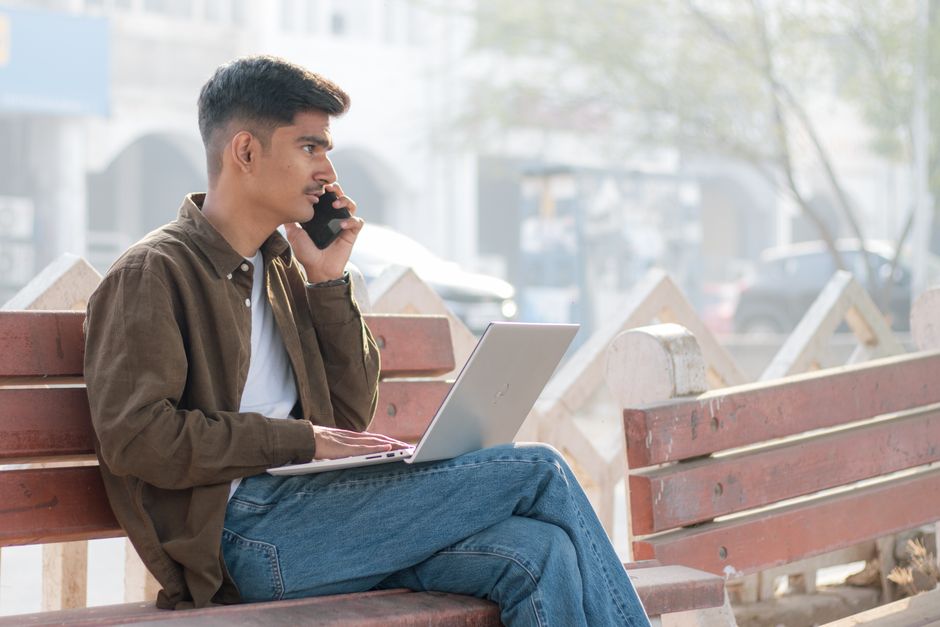 A young man multitasking with a laptop and smartphone on a park bench, embracing remote work outdoors.