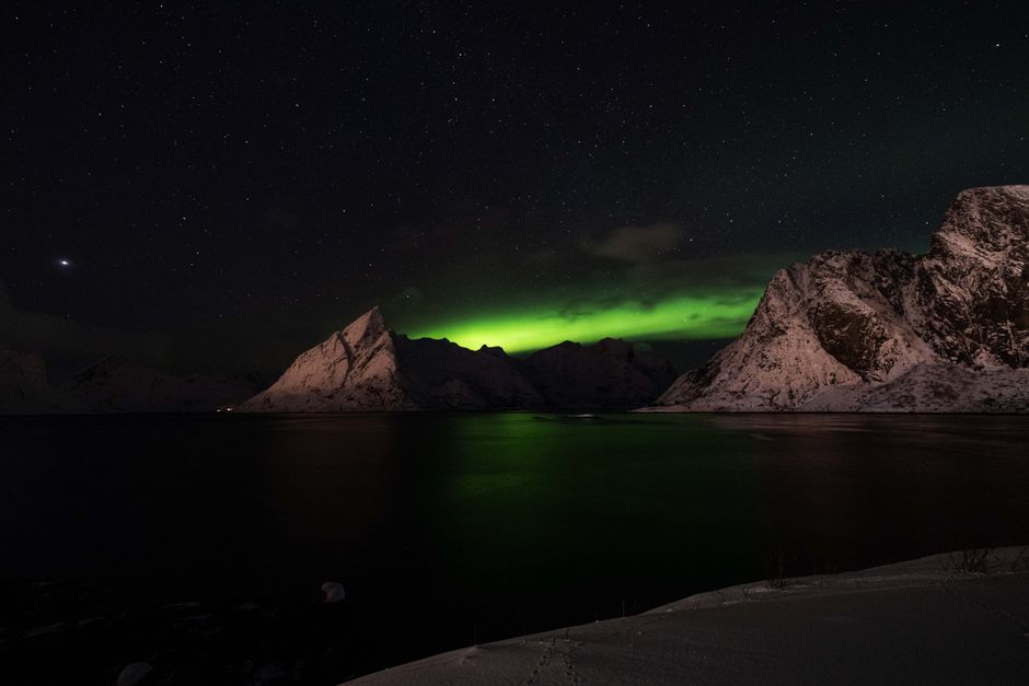 Majestic view of the Northern Lights illuminating snowy mountains and calm waters under a starry sky.