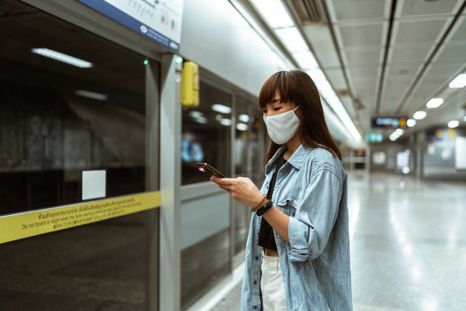 Asian woman wearing a face mask, using her smartphone while waiting at an empty metro station.