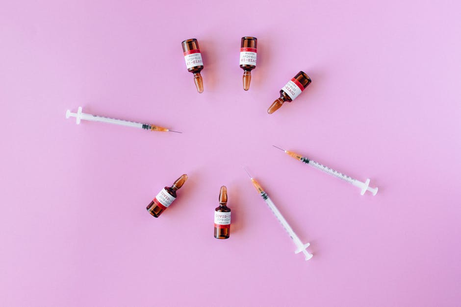 Flat lay of COVID-19 vaccine vials and syringes arranged in a circle on a pink background.