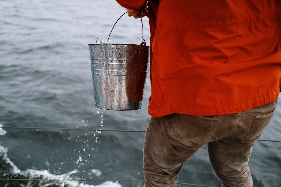 An adult holds a metal bucket on a sailboat at sea, wearing an orange jacket.
