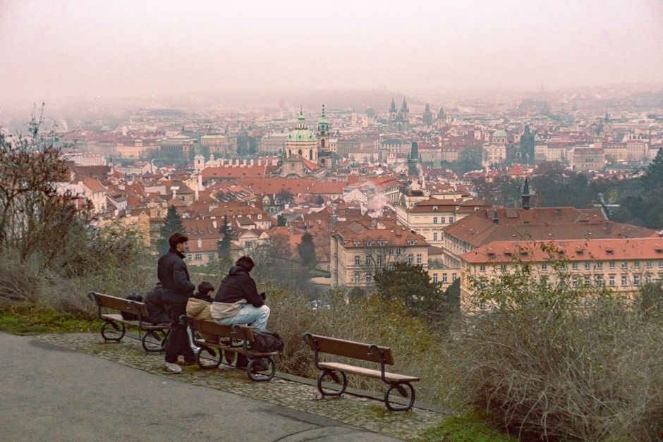 Scenic view of Prague's historic skyline from a hilltop with benches and people enjoying the misty cityscape.